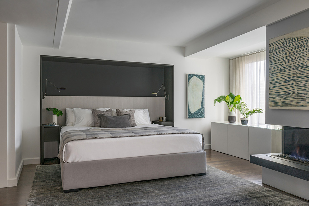 Modelo de dormitorio principal moderno con paredes grises, suelo de madera en tonos medios, chimenea lineal, marco de chimenea de yeso, bandeja y suelo marrón