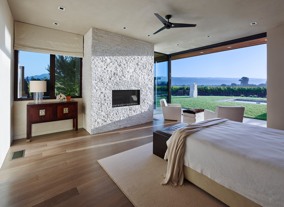 Ejemplo de dormitorio principal moderno de tamaño medio con paredes beige, suelo de madera clara, chimenea lineal y marco de chimenea de piedra