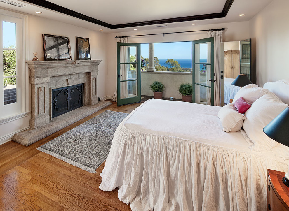 Inspiration for a large mediterranean master bedroom in Santa Barbara with white walls, light hardwood flooring, a standard fireplace, a stone fireplace surround and brown floors.