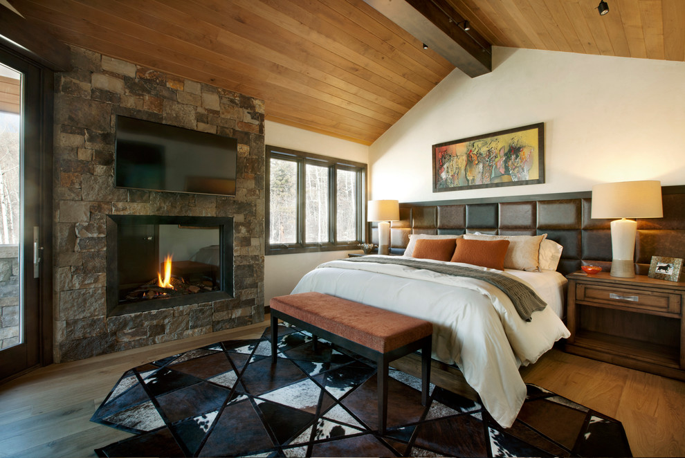 Cette image montre une chambre parentale chalet avec un mur blanc, un sol en bois brun, une cheminée standard, un manteau de cheminée en pierre et un sol marron.