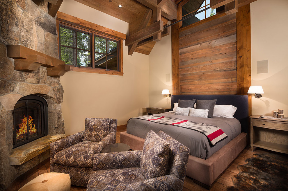 Foto de dormitorio principal rural grande con paredes beige, suelo de madera oscura, chimenea de esquina y marco de chimenea de piedra