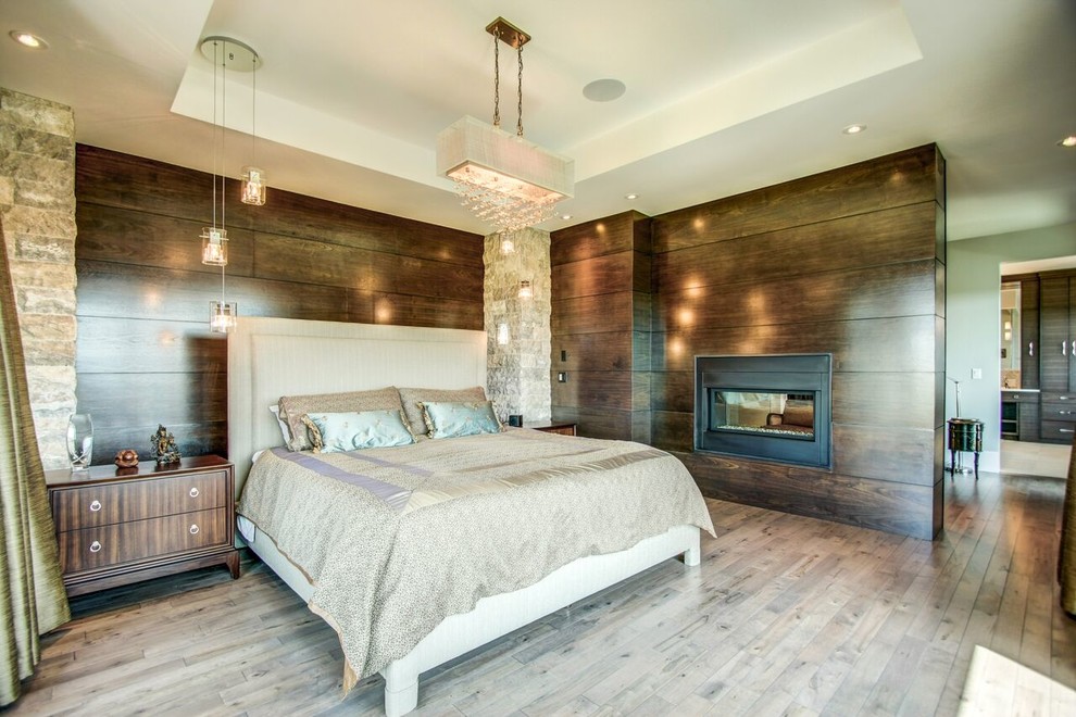 На фото: большая хозяйская спальня в современном стиле с коричневыми стенами, светлым паркетным полом, двусторонним камином и фасадом камина из дерева с