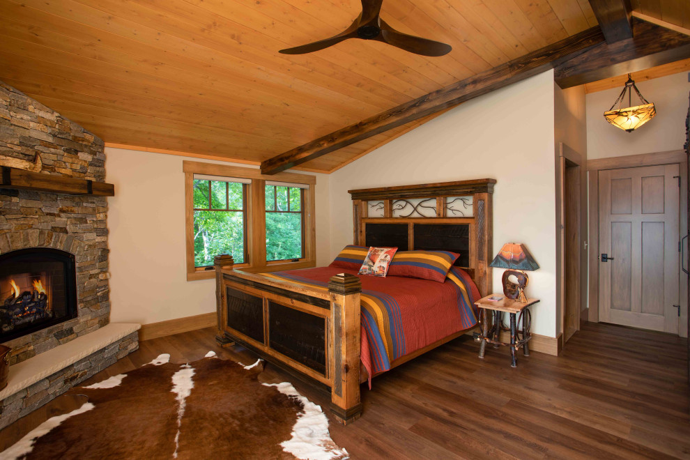 Foto de dormitorio principal rural grande con paredes beige, suelo de madera en tonos medios, chimenea de esquina, marco de chimenea de piedra, suelo marrón y vigas vistas