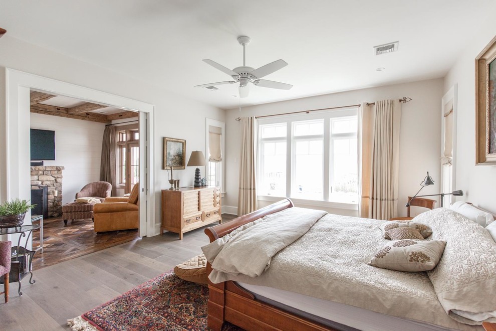 Großes Country Hauptschlafzimmer mit weißer Wandfarbe, braunem Holzboden, Kamin, Kaminumrandung aus Stein und braunem Boden in New York