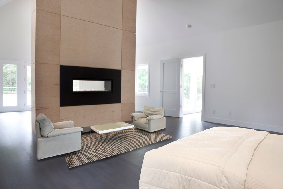 Modelo de dormitorio principal contemporáneo con paredes blancas, suelo de madera oscura, chimenea lineal y marco de chimenea de madera