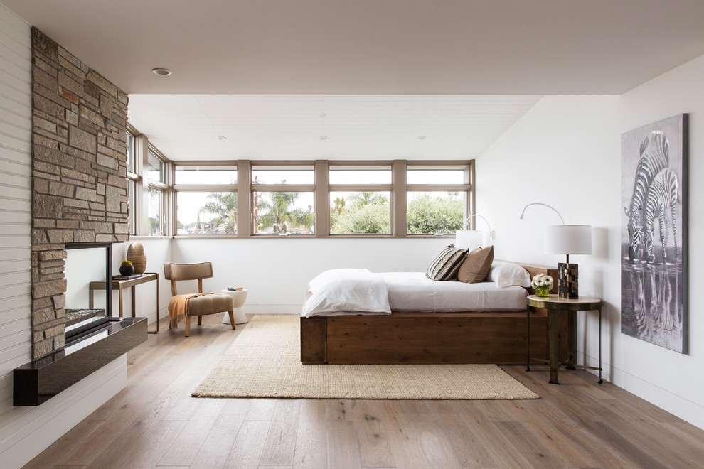 Imagen de dormitorio principal costero de tamaño medio con paredes blancas, suelo de madera en tonos medios, chimenea de doble cara y suelo marrón