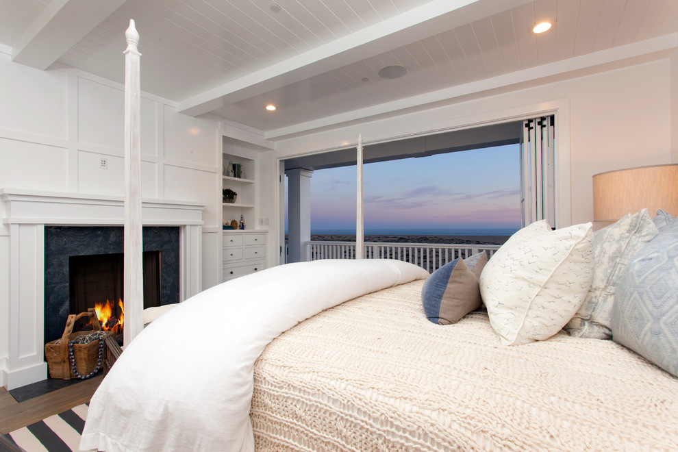 Großes Maritimes Hauptschlafzimmer mit weißer Wandfarbe, braunem Holzboden, Kamin und Kaminumrandung aus Stein in Orange County