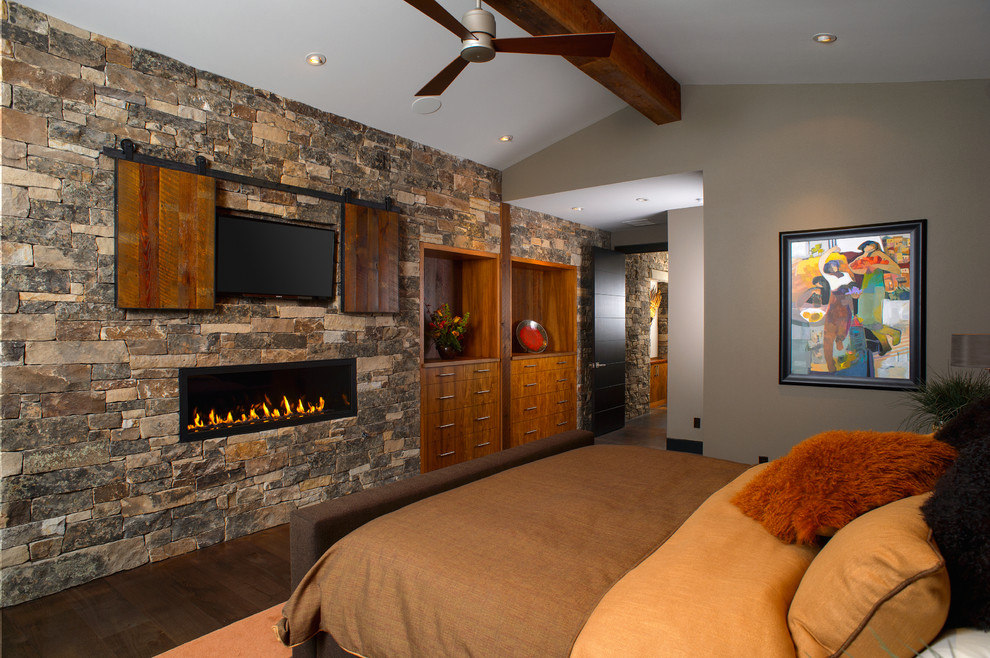 Diseño de dormitorio televisión rural con paredes grises, suelo de madera oscura, chimenea lineal y marco de chimenea de piedra
