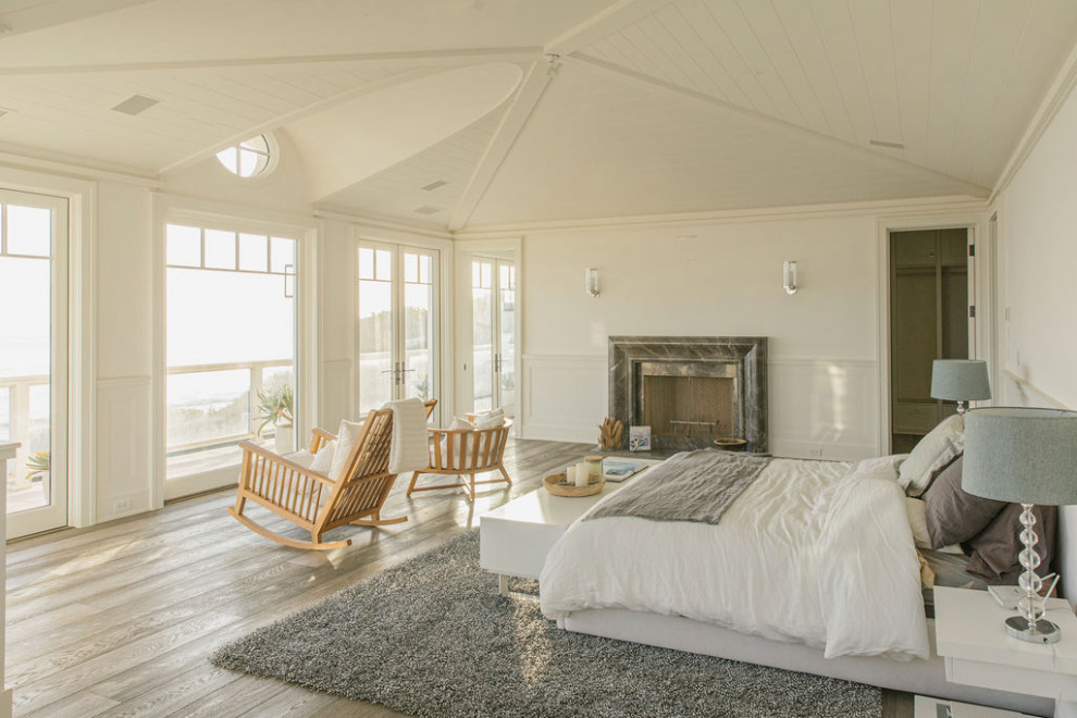 Inspiration for an expansive beach style master bedroom in Los Angeles with white walls, light hardwood flooring, a standard fireplace, a stone fireplace surround, grey floors, a vaulted ceiling and wainscoting.