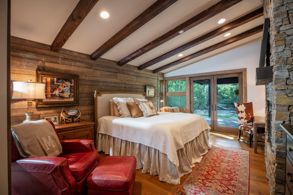 Design ideas for a rustic bedroom in Denver with white walls, medium hardwood flooring, a standard fireplace, a stacked stone fireplace surround, brown floors, exposed beams, a vaulted ceiling and wood walls.