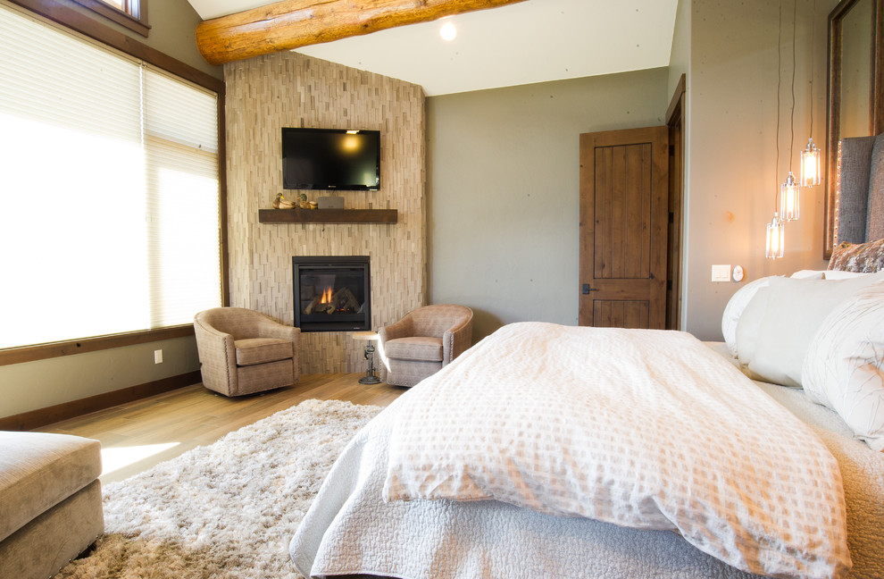 Ejemplo de dormitorio principal rural grande con suelo de baldosas de porcelana, chimenea de esquina, marco de chimenea de piedra, paredes grises y suelo marrón