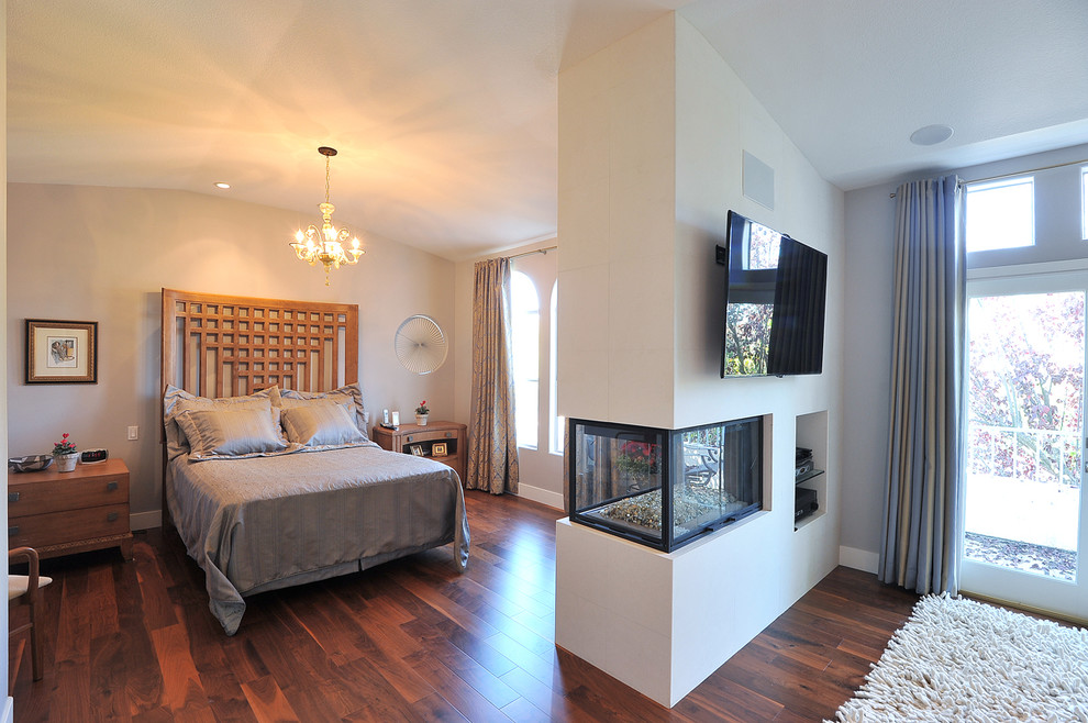 Modelo de dormitorio principal y televisión actual con paredes beige, suelo de madera oscura y chimenea de doble cara