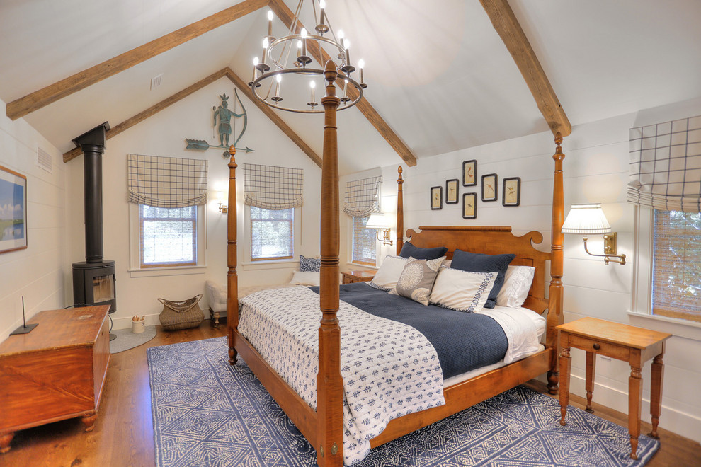 Modelo de dormitorio principal campestre de tamaño medio con paredes blancas, suelo de madera en tonos medios, estufa de leña y suelo marrón