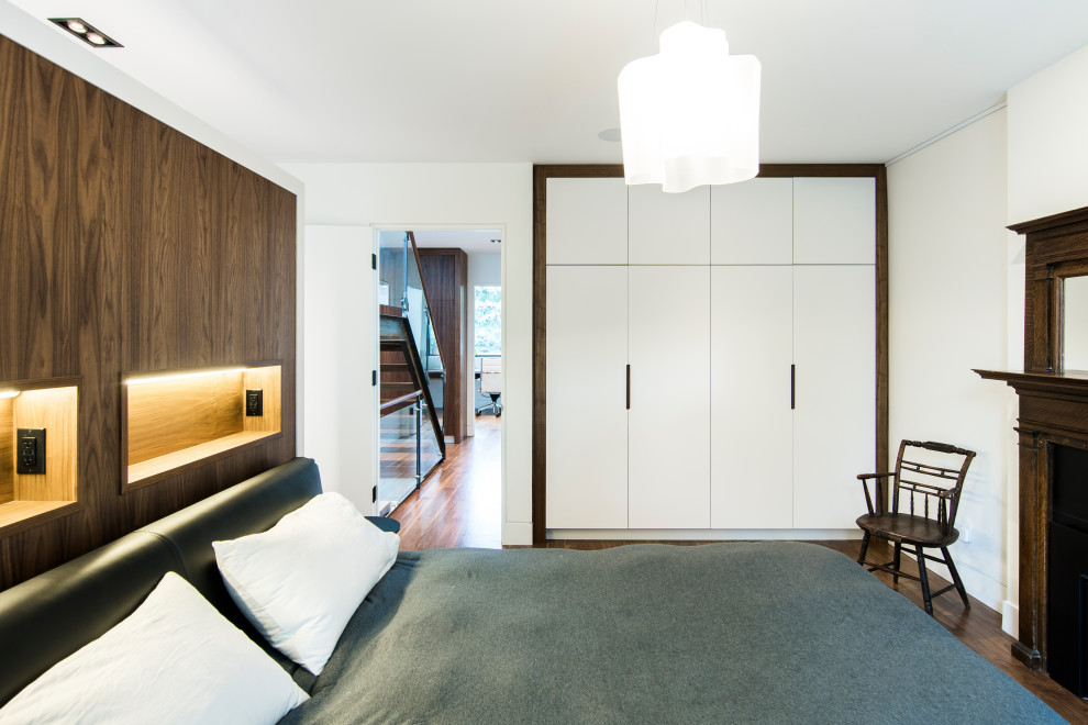 На фото: хозяйская спальня среднего размера в стиле модернизм с белыми стенами, паркетным полом среднего тона, стандартным камином, фасадом камина из дерева и коричневым полом