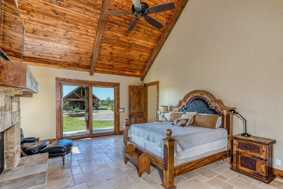 Diseño de dormitorio principal rural grande con paredes beige, suelo de travertino, estufa de leña, marco de chimenea de piedra y suelo beige