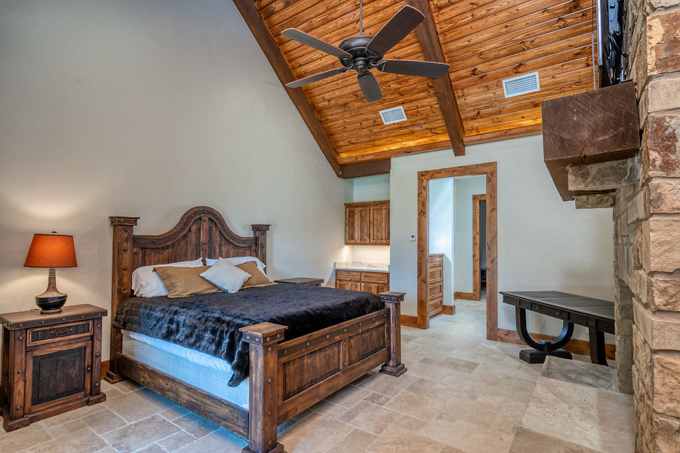 Diseño de dormitorio principal rústico grande con paredes beige, suelo de travertino, estufa de leña, marco de chimenea de piedra y suelo beige