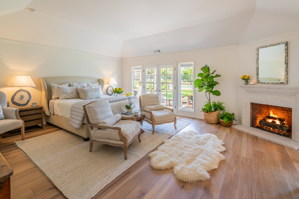 Großes Country Hauptschlafzimmer mit weißer Wandfarbe, braunem Holzboden, Kamin, Kaminumrandung aus Stein, gewölbter Decke und braunem Boden in Santa Barbara
