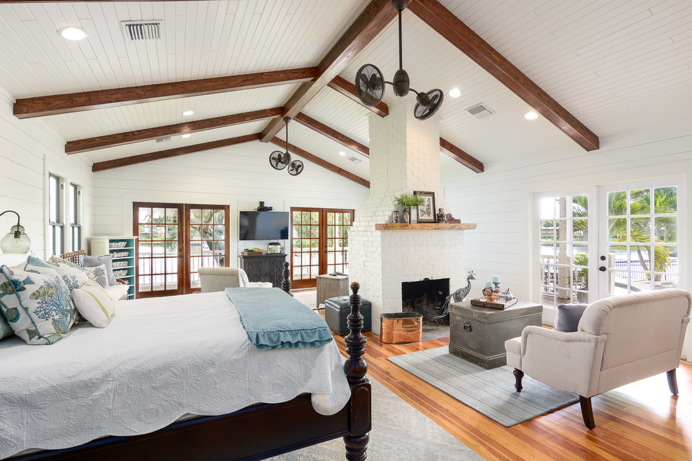Imagen de dormitorio principal costero grande con paredes blancas, marco de chimenea de ladrillo, suelo de madera en tonos medios, todas las chimeneas y suelo marrón