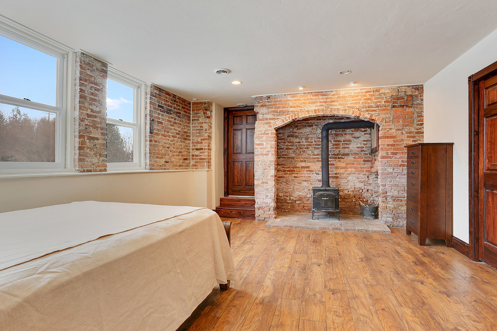 Foto de habitación de invitados rural grande con paredes blancas, suelo de madera en tonos medios, estufa de leña y marco de chimenea de ladrillo