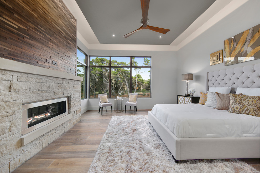 Großes Klassisches Hauptschlafzimmer mit grauer Wandfarbe, hellem Holzboden, Gaskamin, Kaminumrandung aus Stein und braunem Boden in Austin