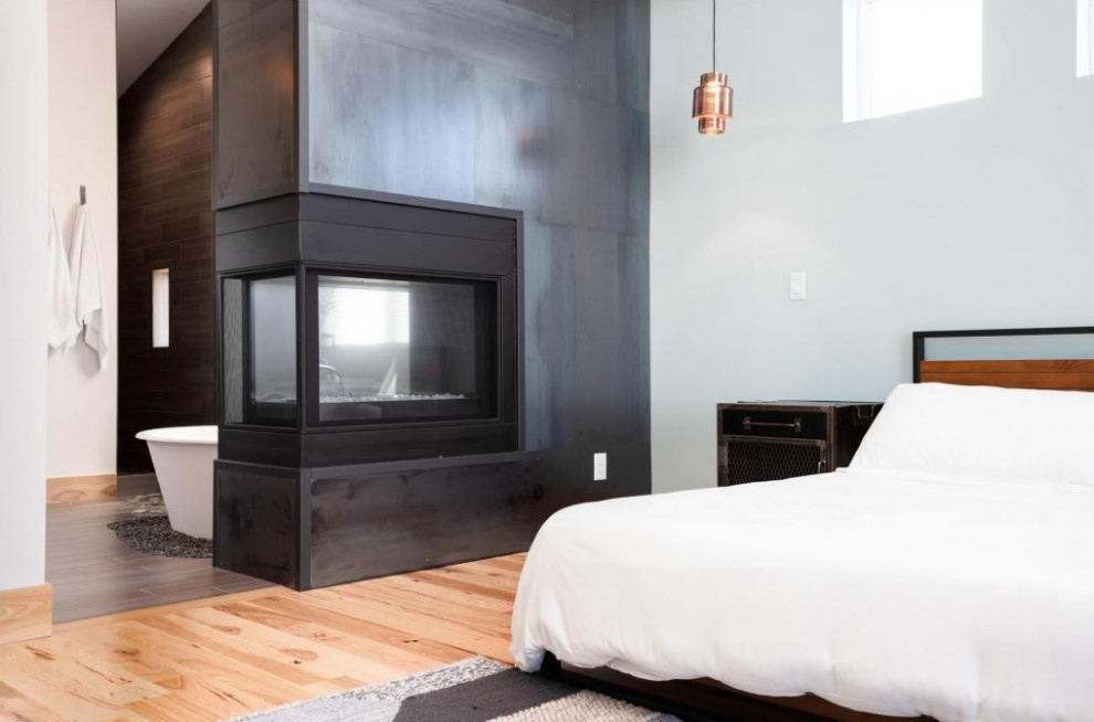Modelo de dormitorio urbano grande con paredes grises, suelo de madera clara, chimenea de doble cara y marco de chimenea de metal