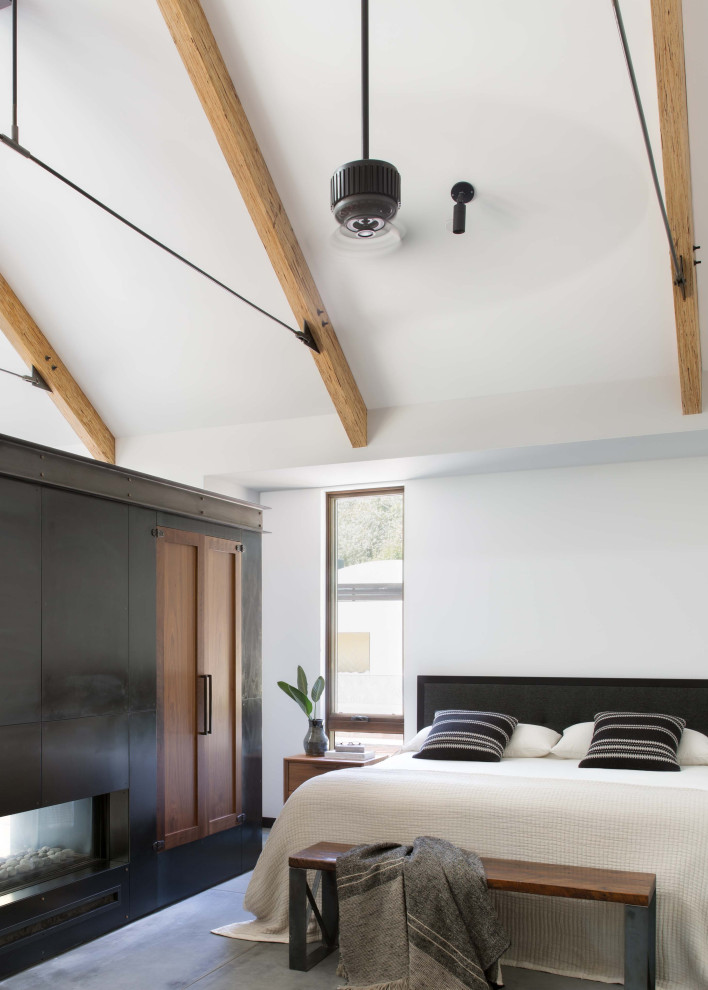 Farmhouse master bedroom in San Francisco with a two-sided fireplace, a metal fireplace surround, white walls, concrete flooring, grey floors, exposed beams and a vaulted ceiling.