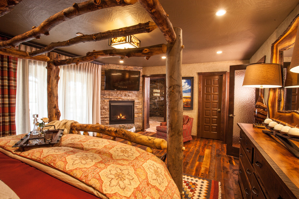 Ejemplo de dormitorio principal rural grande con paredes beige, suelo de madera en tonos medios, chimenea de esquina y marco de chimenea de piedra