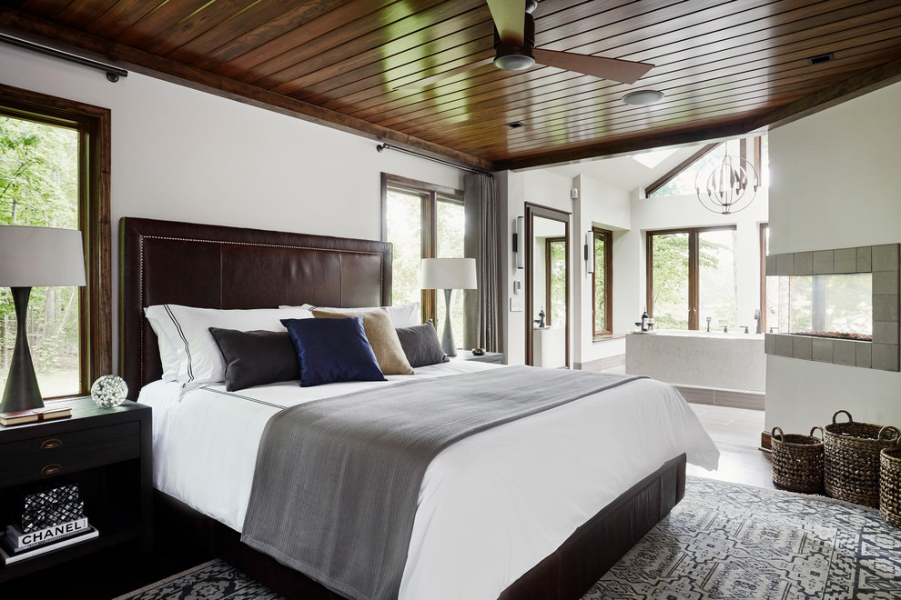 Modelo de dormitorio principal minimalista grande con paredes blancas, suelo de madera oscura, chimenea de doble cara y marco de chimenea de baldosas y/o azulejos