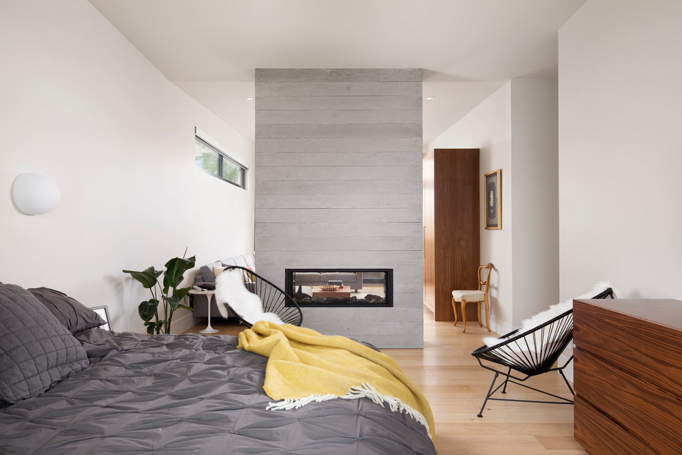 Modernes Hauptschlafzimmer mit weißer Wandfarbe, hellem Holzboden, Kaminumrandung aus Beton, Tunnelkamin und beigem Boden in Calgary