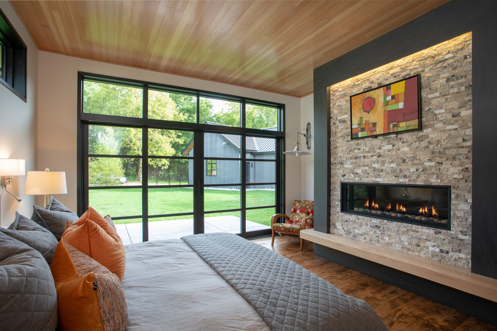 Cette photo montre une grande chambre parentale moderne avec un mur blanc, un sol en bois brun, une cheminée ribbon, un manteau de cheminée en pierre et un sol marron.