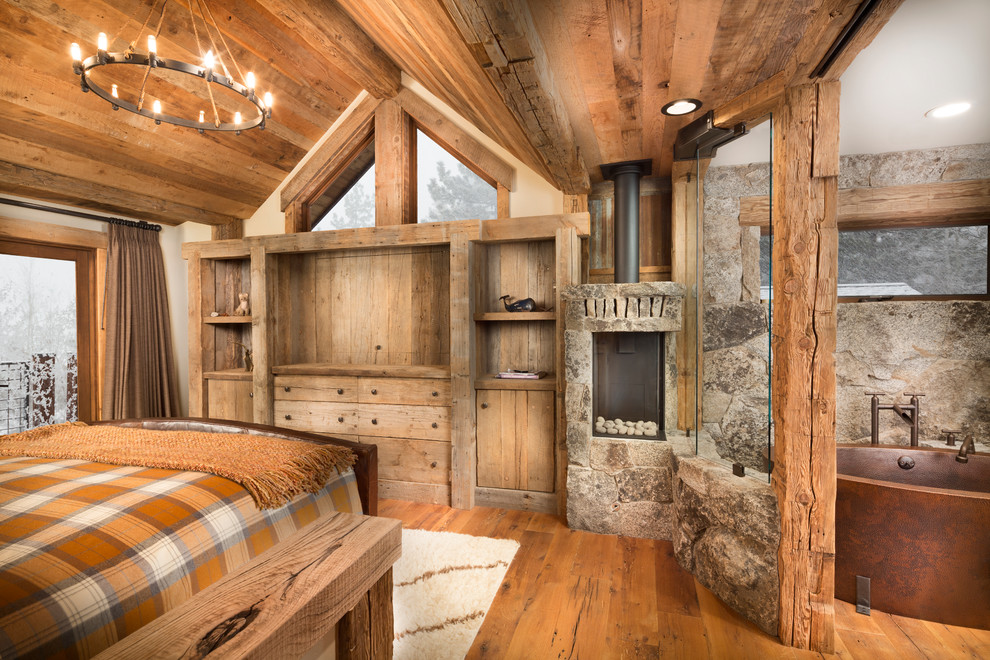 Idée de décoration pour une chambre parentale chalet de taille moyenne avec un mur beige, un sol en bois brun, un manteau de cheminée en pierre, un poêle à bois et un sol orange.