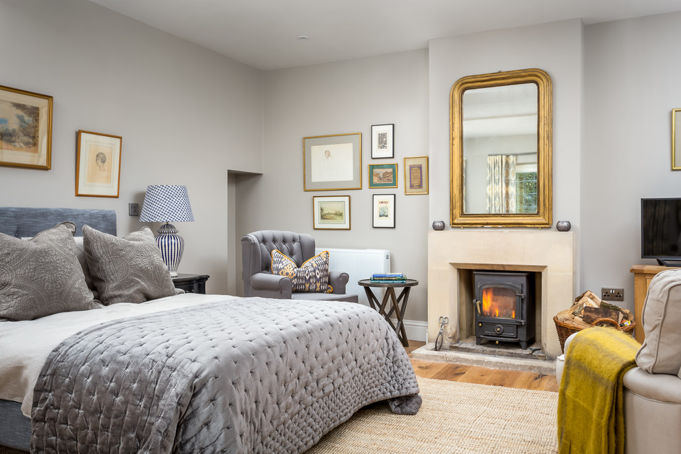 グロスタシャーにある広いカントリー風のおしゃれな主寝室 (グレーの壁、無垢フローリング、薪ストーブ、石材の暖炉まわり)