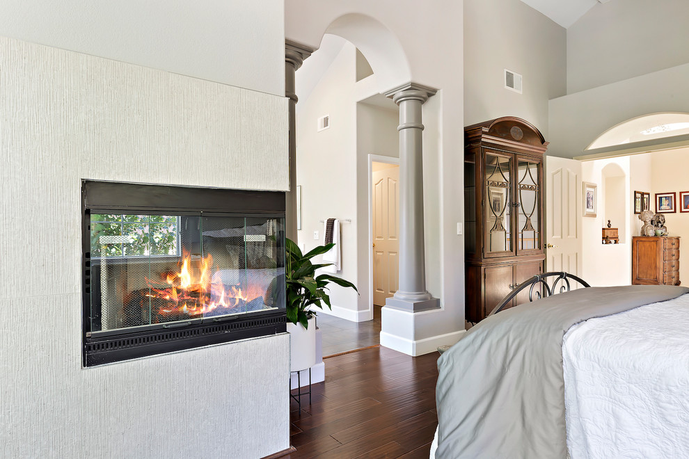 Photo of a large contemporary master bedroom in Other with beige walls, dark hardwood flooring, a two-sided fireplace, a tiled fireplace surround and brown floors.