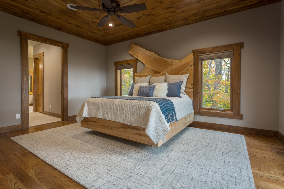 Modelo de dormitorio principal rústico grande sin chimenea con paredes grises, suelo de madera en tonos medios y suelo marrón