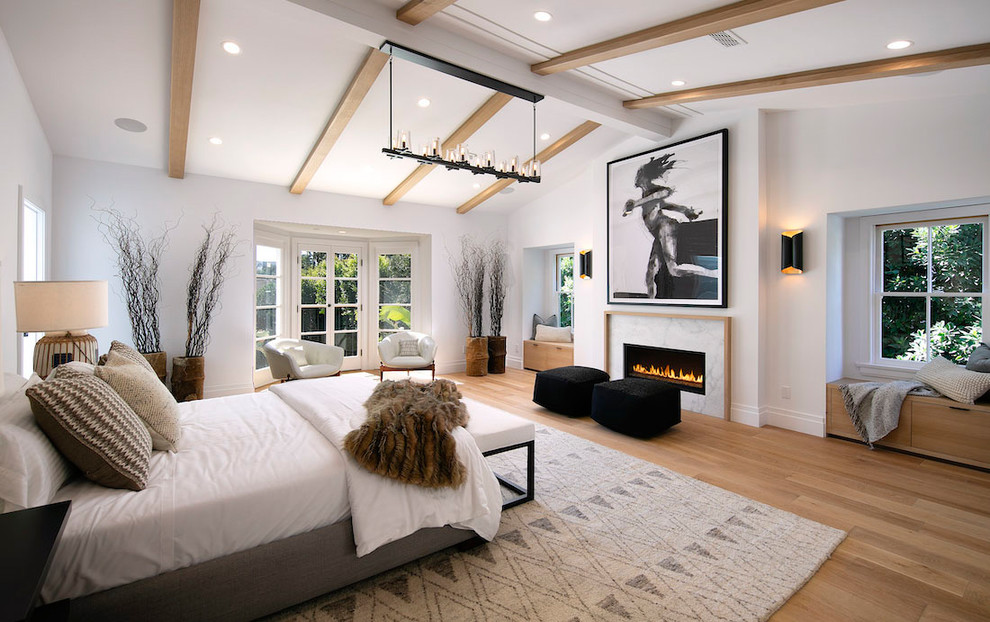 Imagen de dormitorio principal costero con paredes blancas, suelo de madera clara, chimenea lineal y marco de chimenea de piedra
