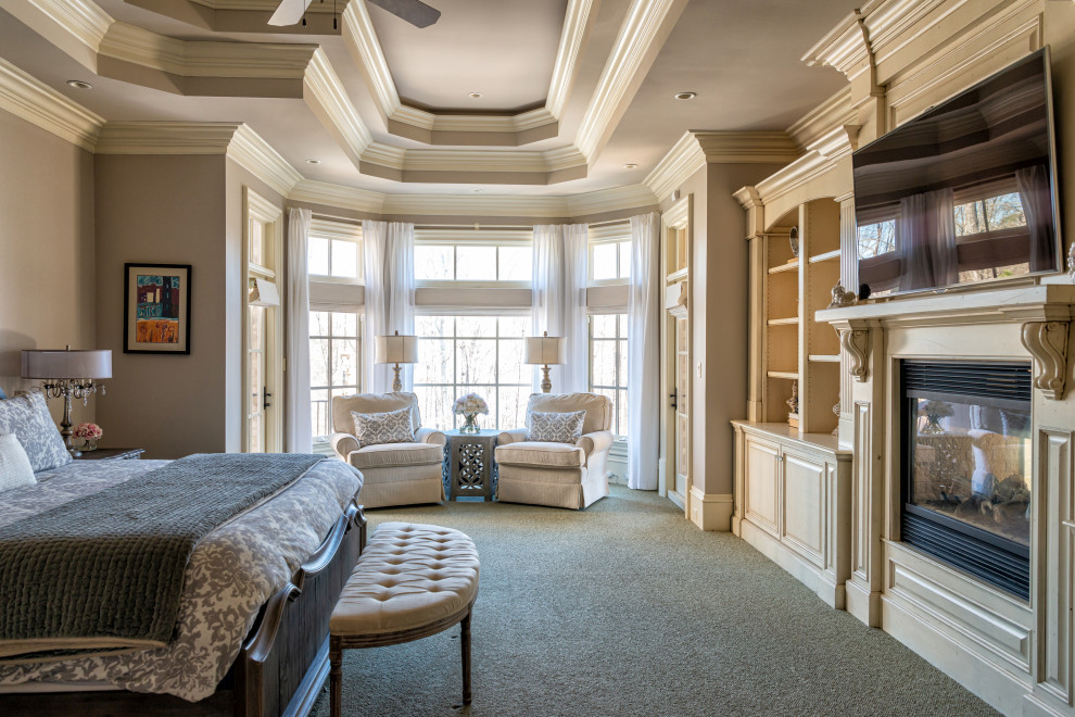 На фото: огромная хозяйская спальня с серыми стенами, ковровым покрытием, двусторонним камином, фасадом камина из дерева, серым полом и многоуровневым потолком