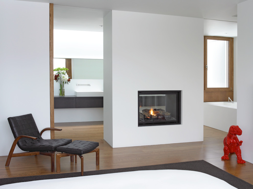 Imagen de dormitorio principal contemporáneo de tamaño medio con paredes blancas, suelo de madera en tonos medios, chimenea de doble cara y marco de chimenea de yeso