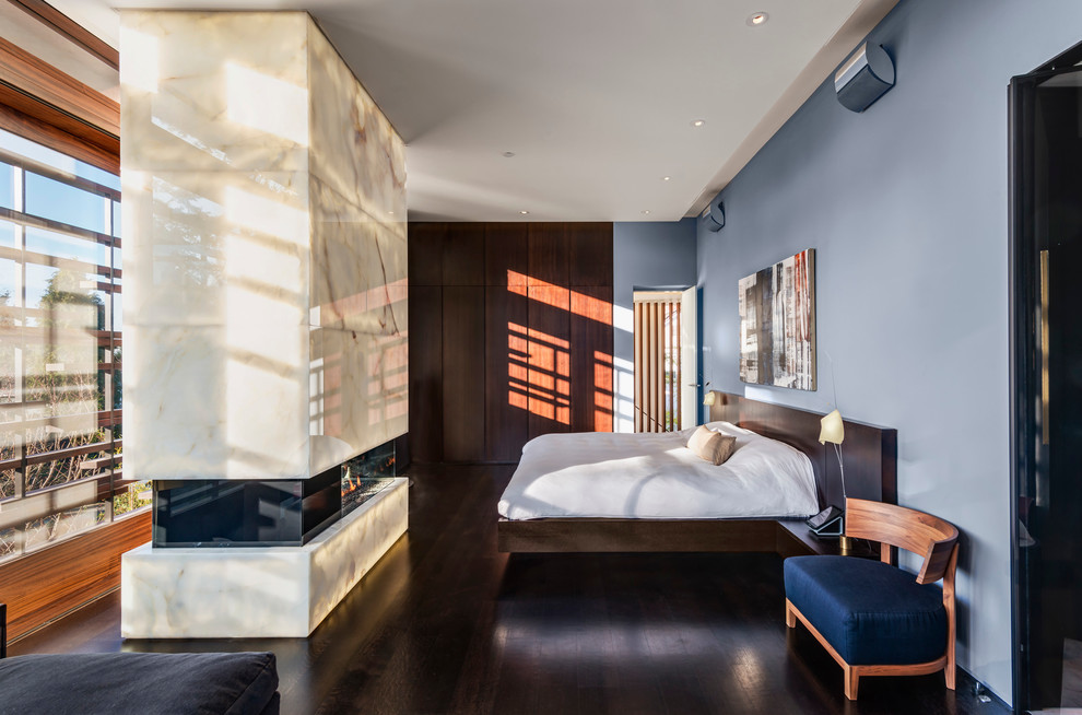 Mittelgroßes Modernes Hauptschlafzimmer mit blauer Wandfarbe, dunklem Holzboden, Tunnelkamin, Kaminumrandung aus Stein und braunem Boden in Vancouver