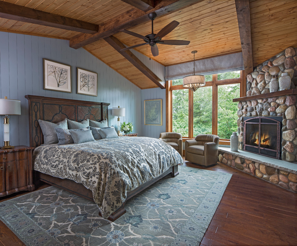 Cette image montre une chambre parentale chalet avec un mur bleu, un sol en bois brun, une cheminée d'angle, un manteau de cheminée en pierre, un sol marron, poutres apparentes et du lambris de bois.