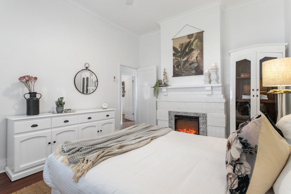 Bild på ett vintage sovrum, med vita väggar, mörkt trägolv, en standard öppen spis, en spiselkrans i tegelsten och brunt golv