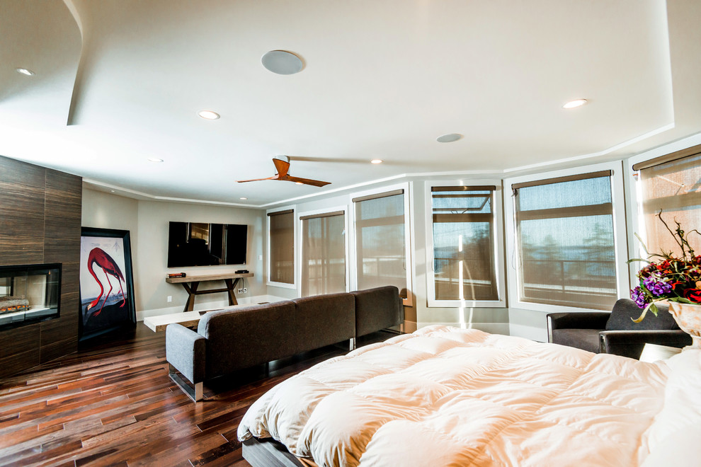 Ejemplo de dormitorio principal minimalista grande con paredes grises, suelo de madera en tonos medios, chimenea de doble cara y marco de chimenea de baldosas y/o azulejos
