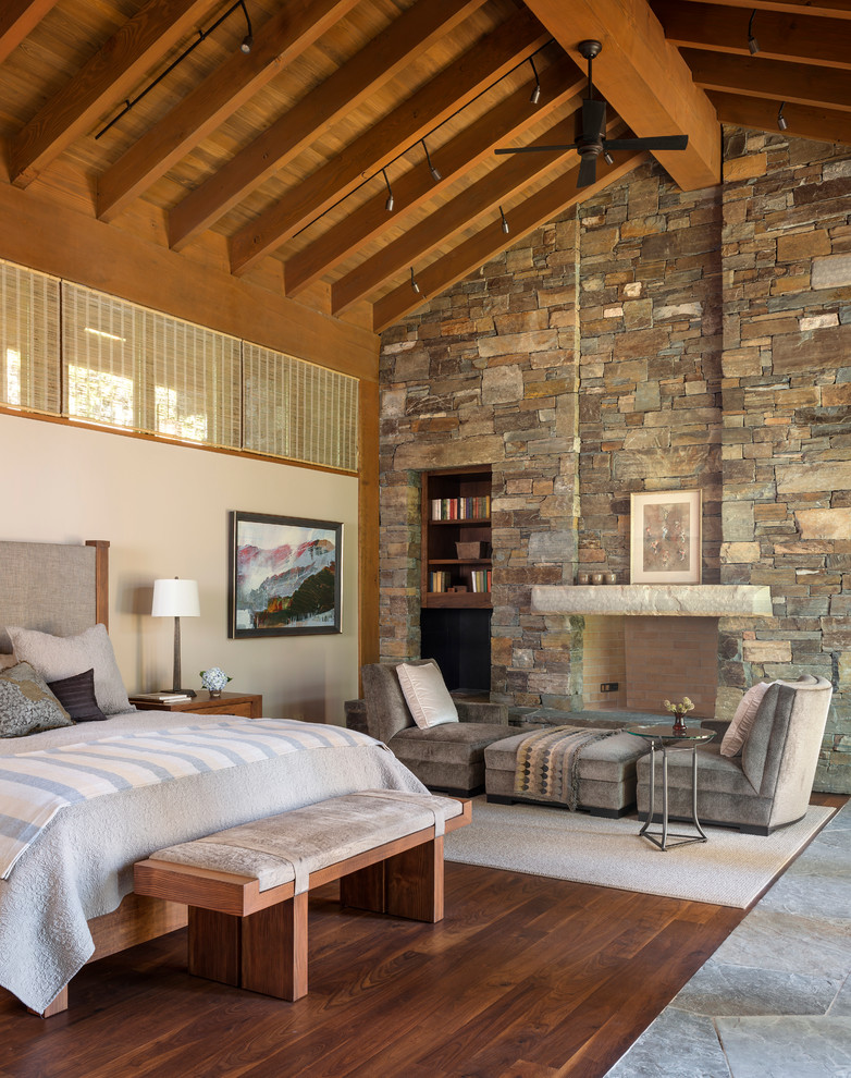 Modelo de dormitorio principal rústico con paredes beige, suelo de madera en tonos medios, todas las chimeneas y marco de chimenea de piedra