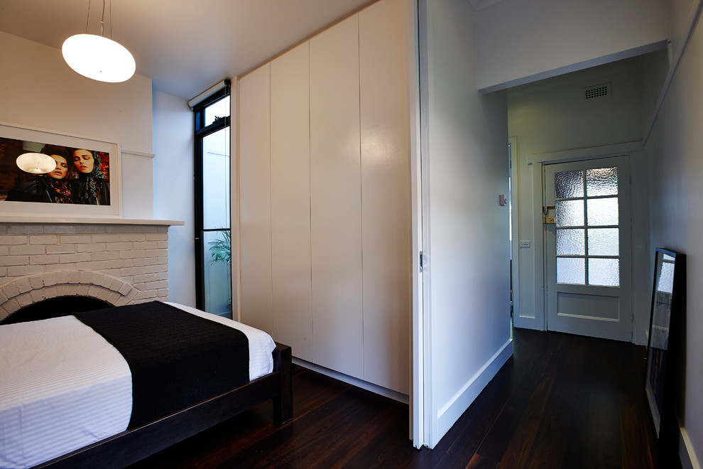 Modelo de dormitorio principal contemporáneo pequeño con paredes blancas, suelo de madera oscura, todas las chimeneas y marco de chimenea de ladrillo