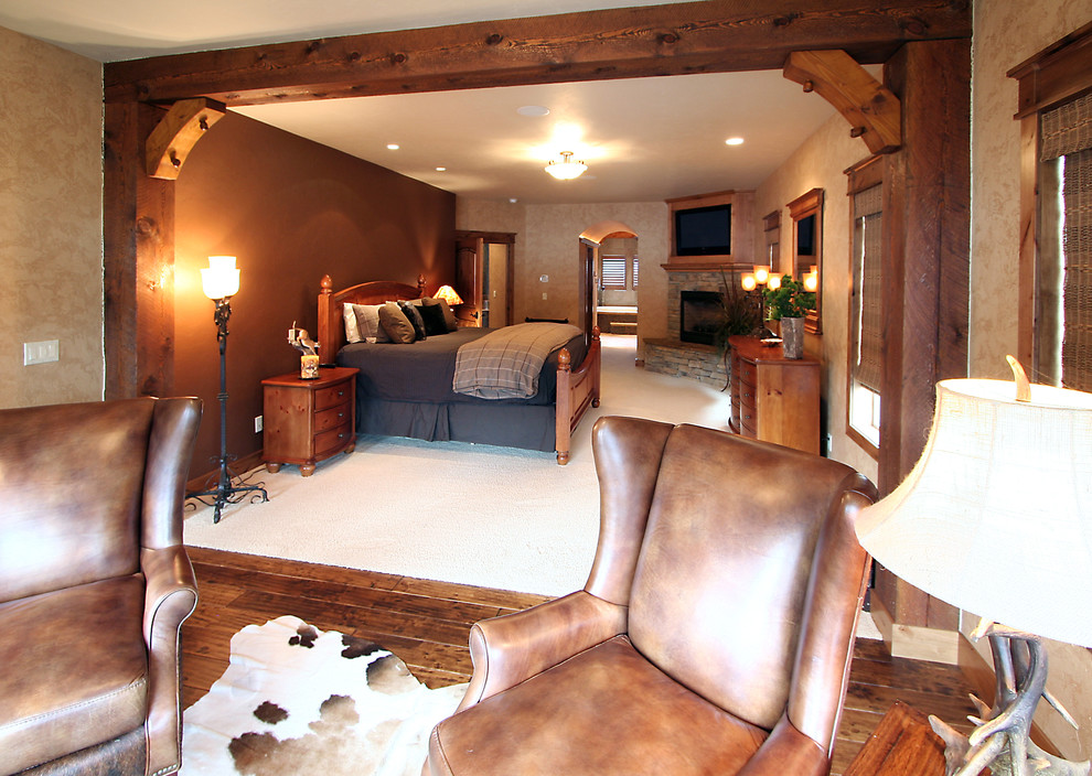 На фото: большая хозяйская спальня в стиле рустика с бежевыми стенами, ковровым покрытием, угловым камином и фасадом камина из камня