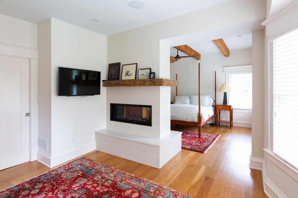 Imagen de dormitorio principal de estilo americano de tamaño medio con paredes blancas, suelo de madera en tonos medios, chimenea de doble cara y marco de chimenea de baldosas y/o azulejos