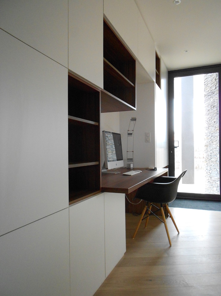 Bild på ett mellanstort funkis hemmabibliotek, med vita väggar, ljust trägolv och ett inbyggt skrivbord