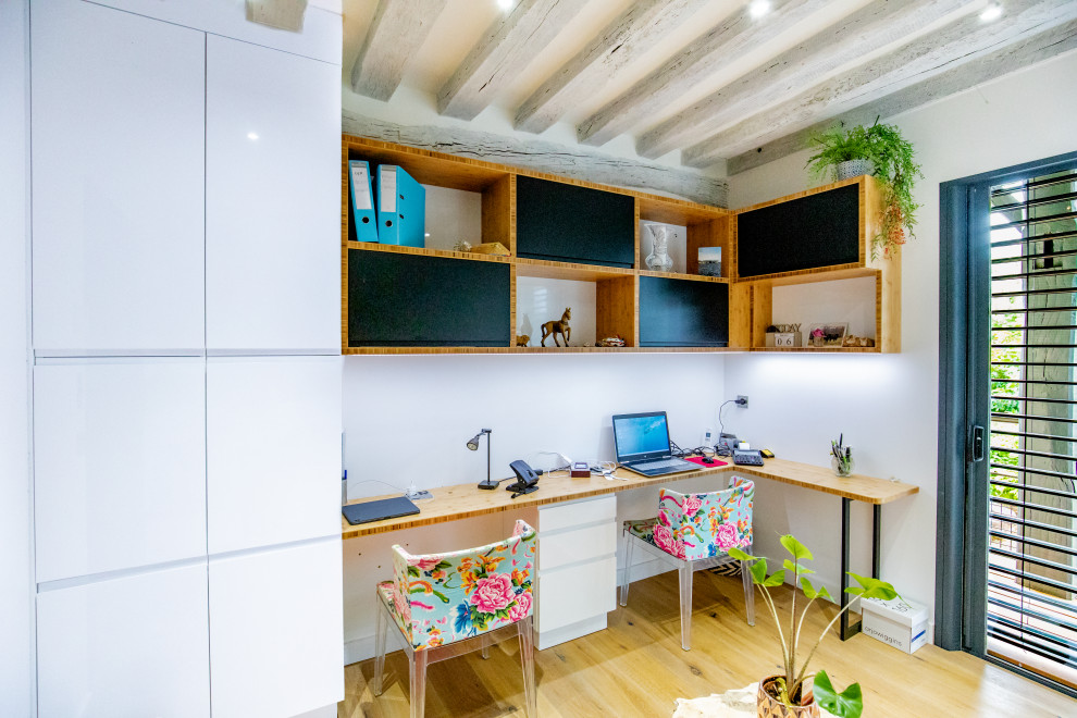 Modelo de despacho bohemio con paredes blancas, suelo de madera en tonos medios, escritorio empotrado, suelo marrón y vigas vistas