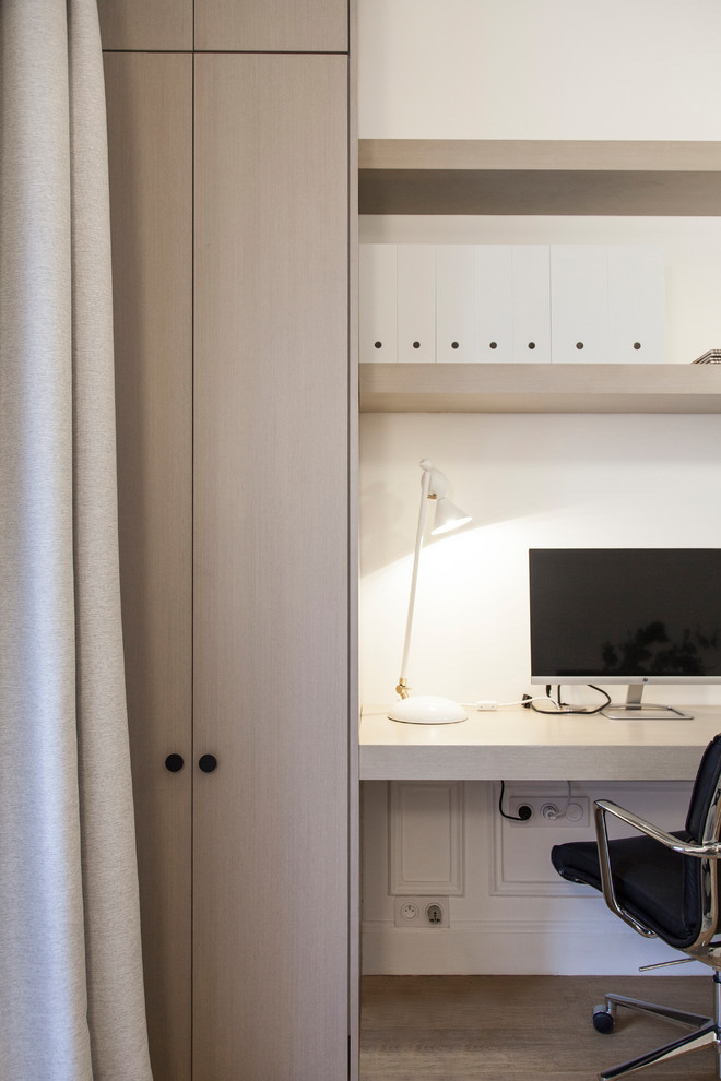 Modelo de despacho actual de tamaño medio sin chimenea con paredes blancas, suelo de madera en tonos medios, escritorio empotrado y suelo marrón