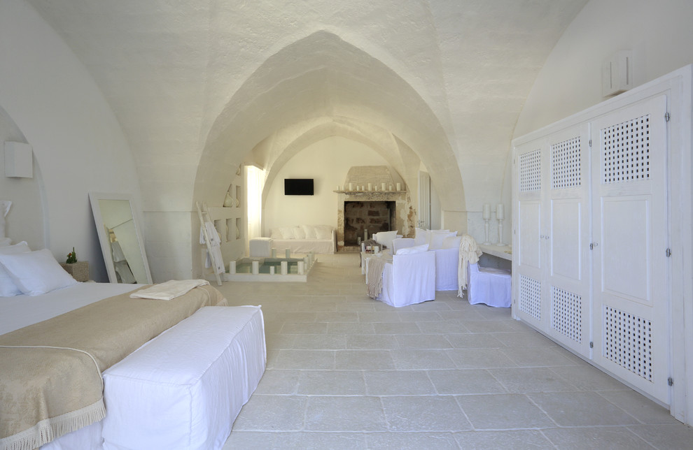 Modelo de dormitorio principal mediterráneo extra grande con paredes blancas, suelo de piedra caliza, todas las chimeneas y marco de chimenea de piedra