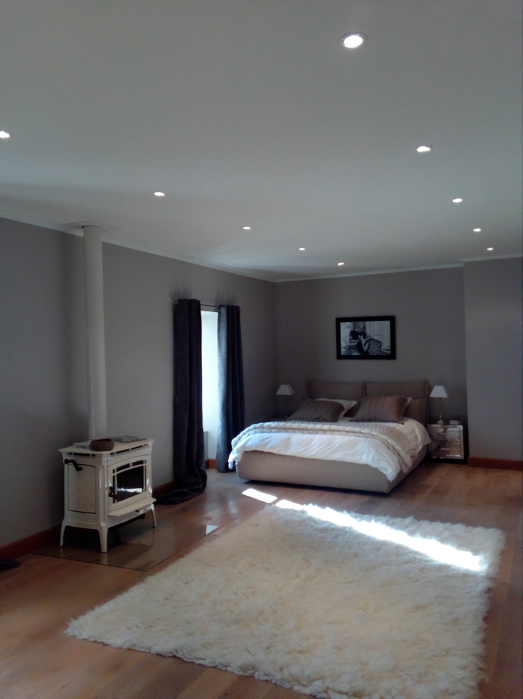 Diseño de dormitorio principal clásico renovado extra grande con paredes grises, suelo de madera clara y estufa de leña